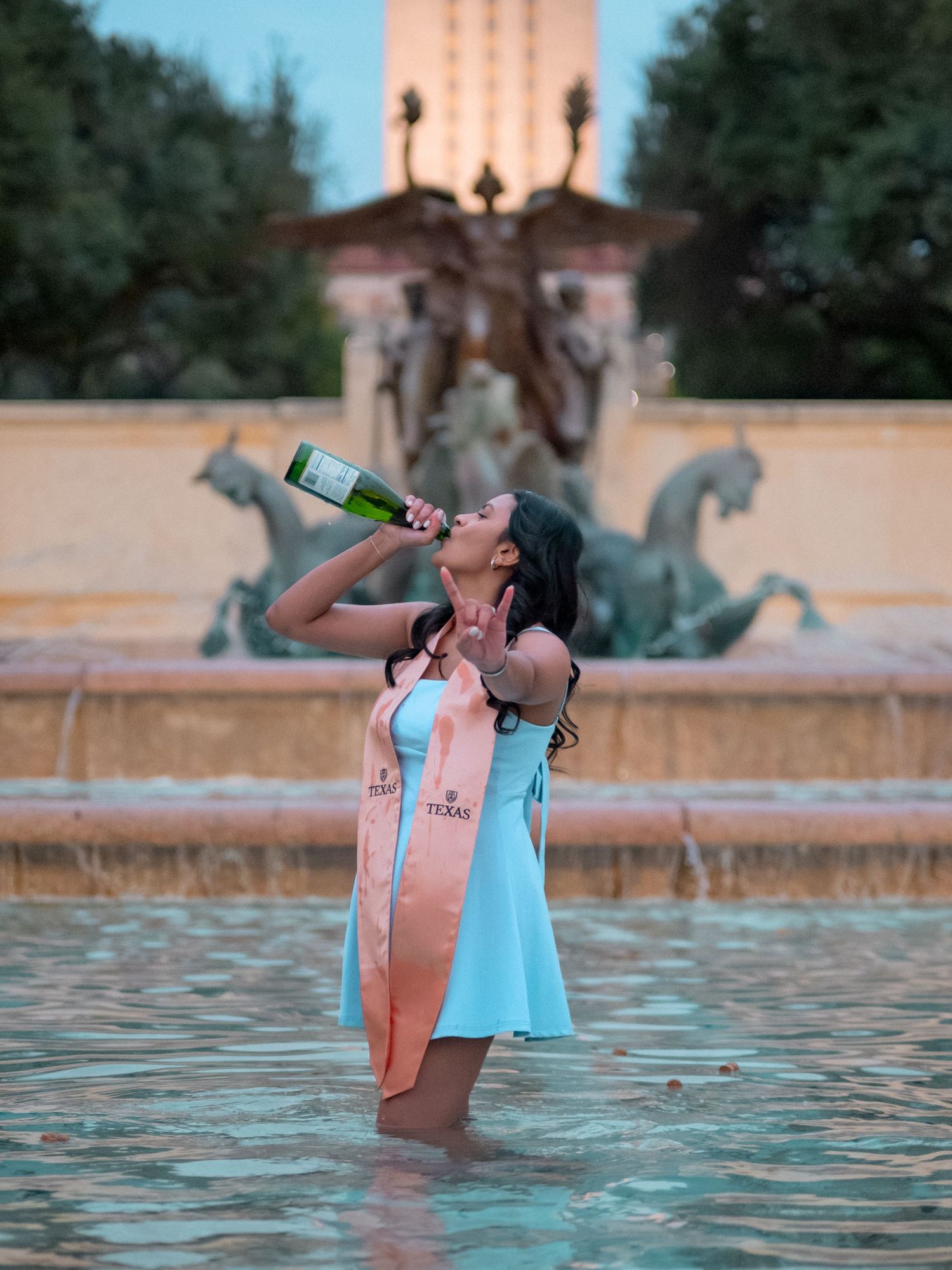 Person in light blue dress stands in fountain at dusk drinking from bottle with ornate statue and architecture in background.