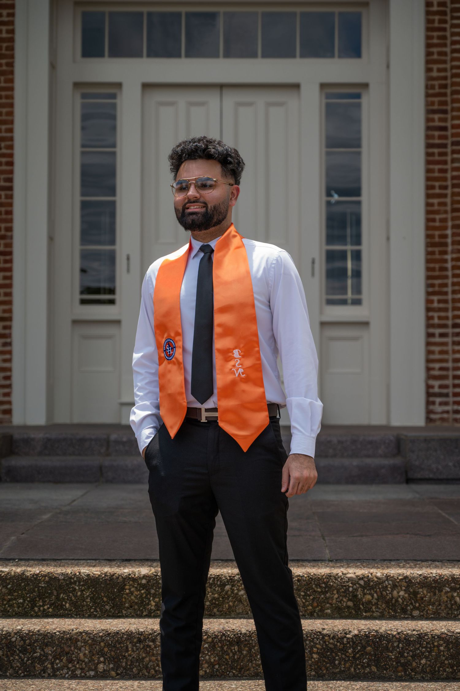 Graduate in white shirt and orange stole poses in front of brick building.