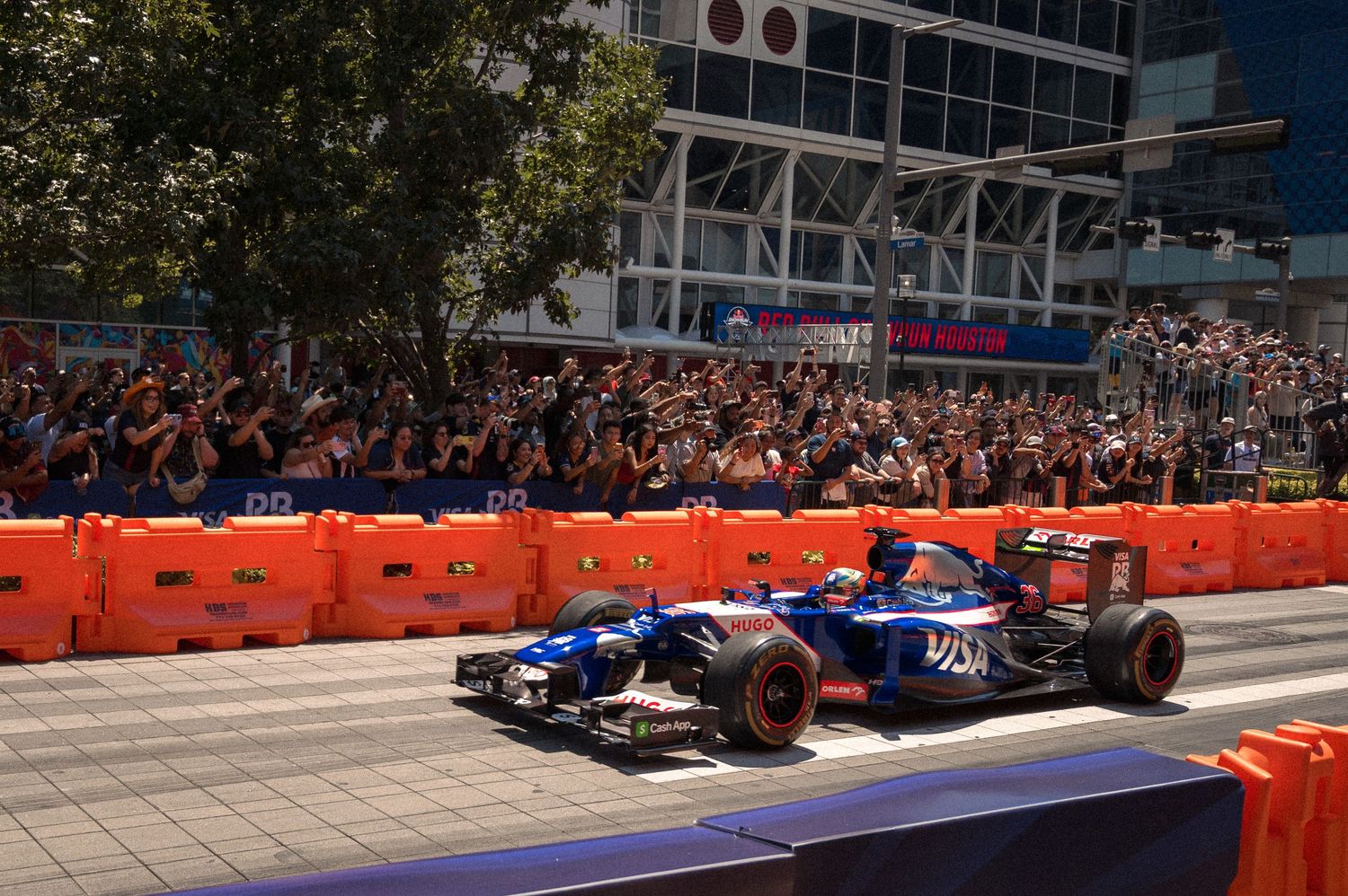 F1 cars race past modern buildings on temporary street circuit with orange safety barriers and packed grandstands.