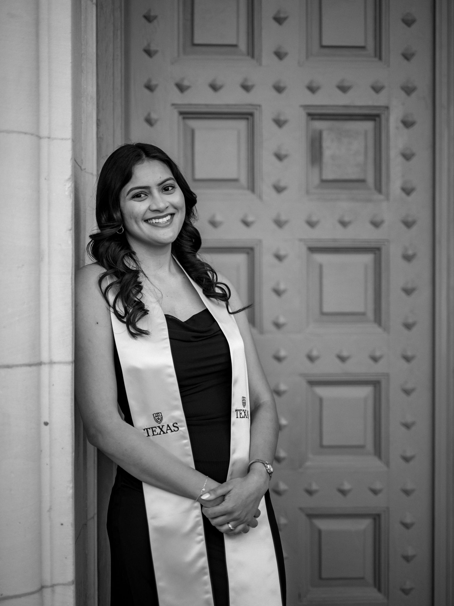 A graduate in a black dress and white graduation stole poses for a portrait in front of a decorative door.