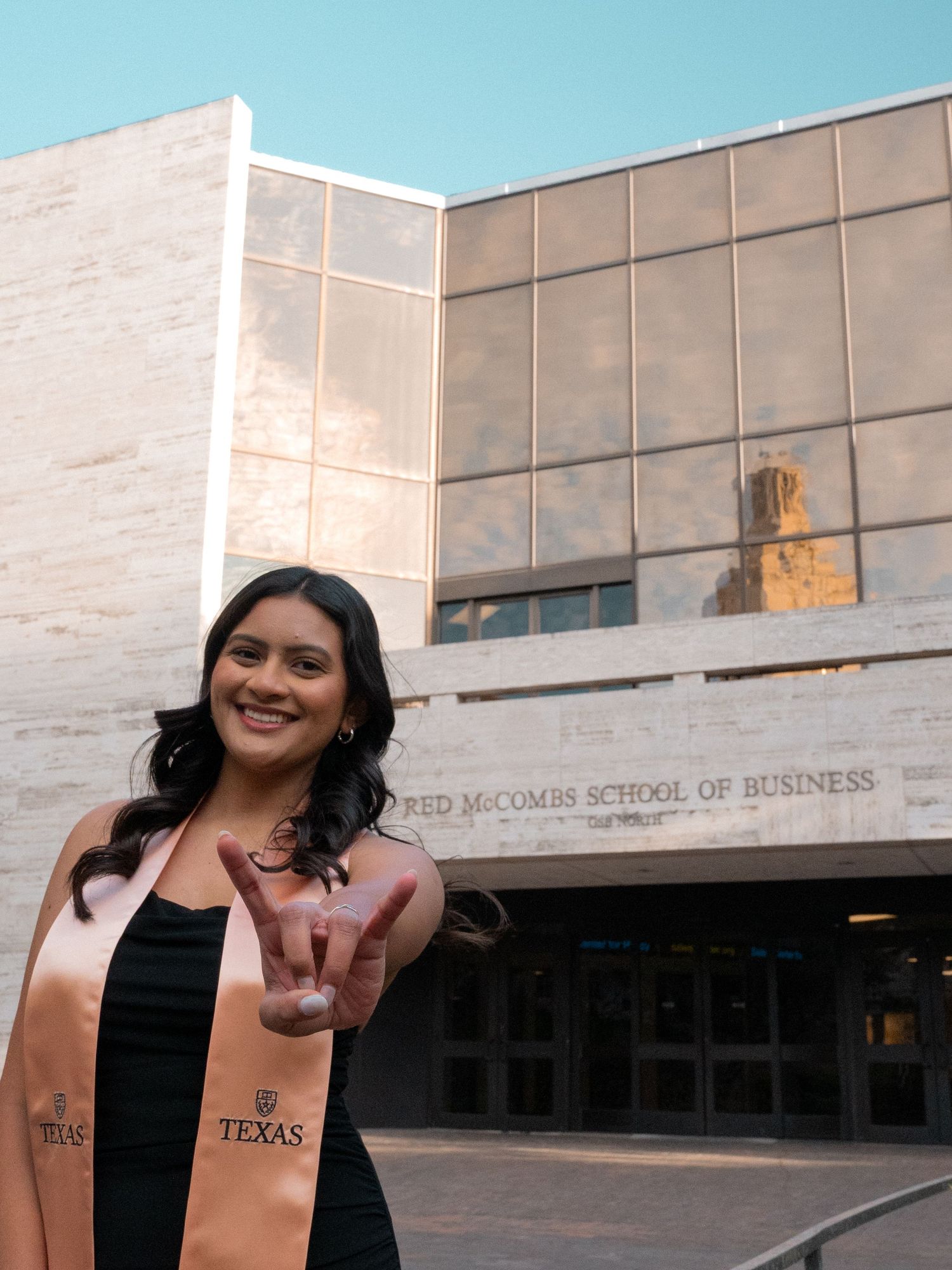 A graduate wearing a salmon-colored graduation stole stands outside McCombs School of Business at the University of Texas.