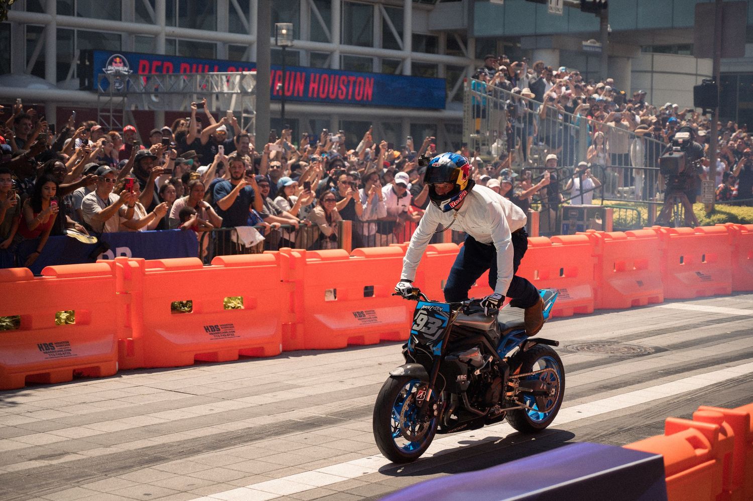 Motorcycle racing demonstration on closed city street with crowd barriers and spectators.