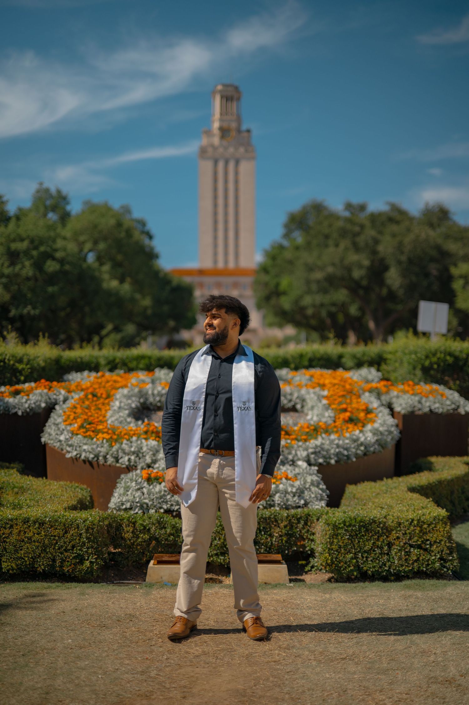 Graduate poses in front of UT Tower with orange and white floral display in foreground.