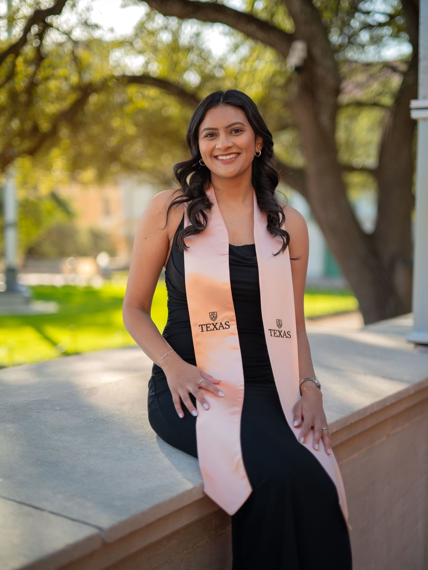 Graduate in black dress and pink stole poses outdoors at Texas university campus during sunny day.