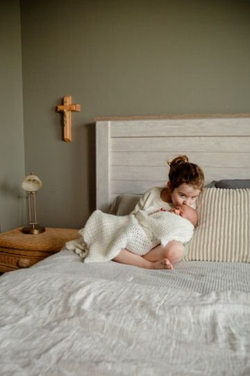 Indoor Christian lifestyle family photography session with sister kissing newborn with Catholic imagery in Omaha, Nebraska