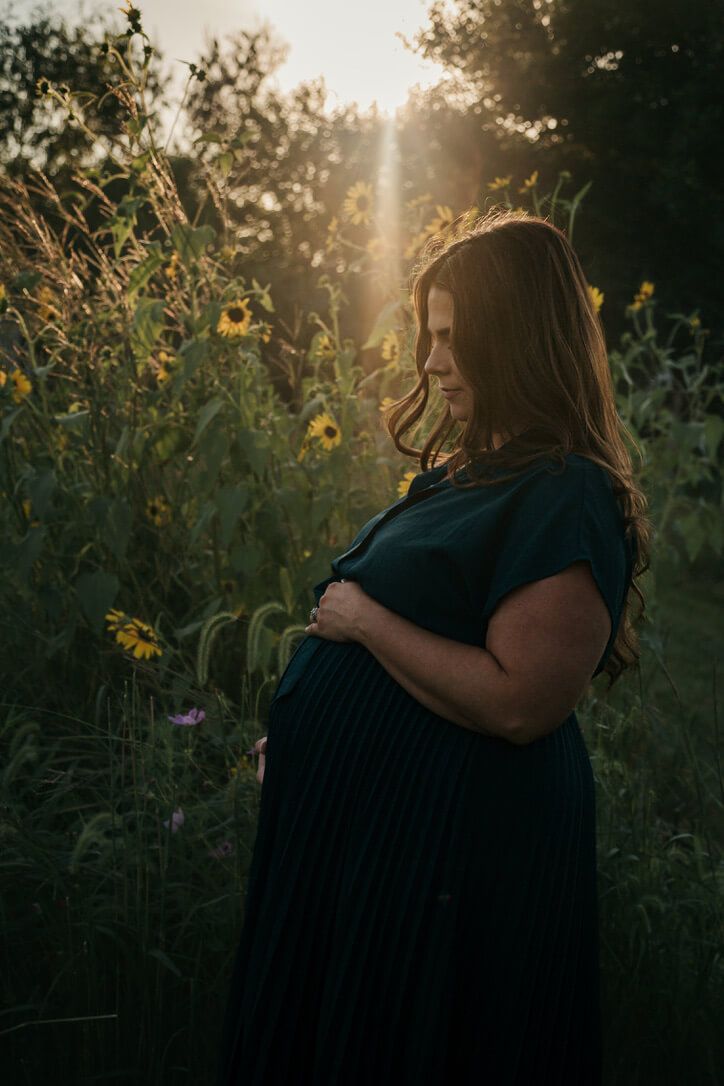 Mother looking at baby bump surrounded by sunflowers from outdoor maternity lifestyle photography session during golden hour in Omaha, NE