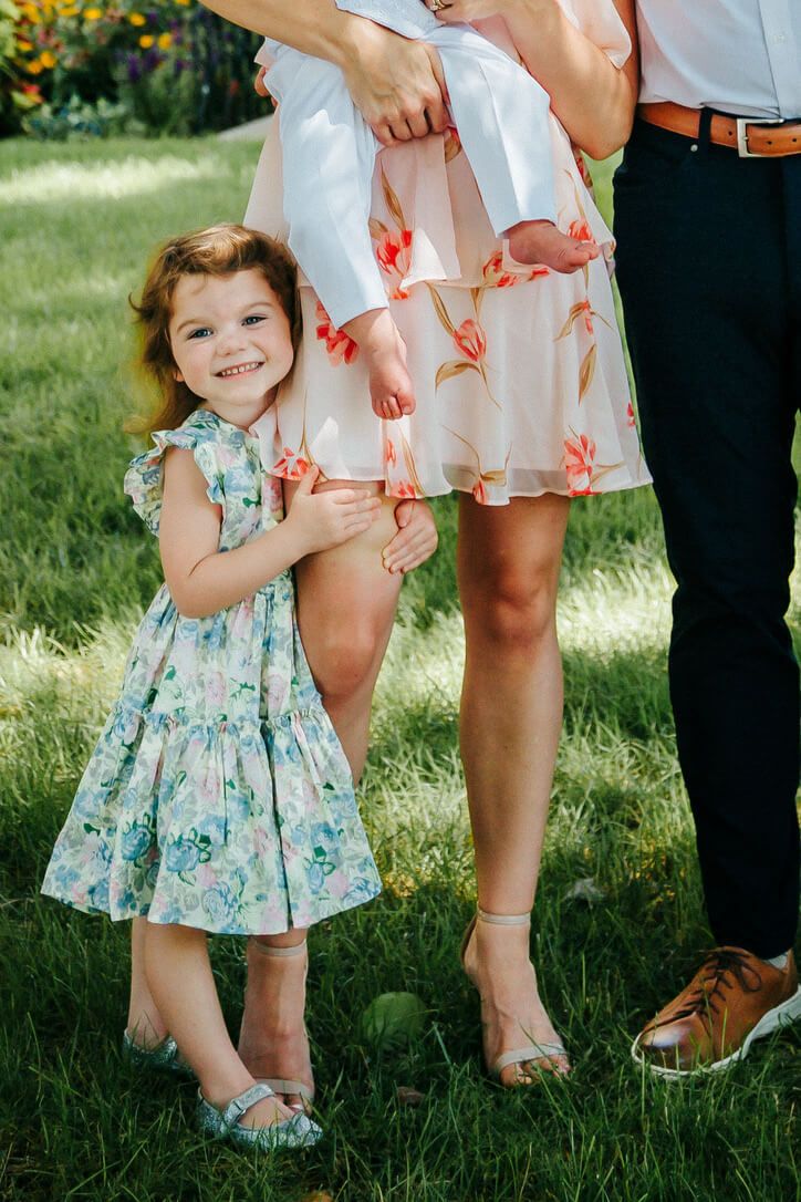 Outdoor family photography session with child holding mother's leg on baby's Catholic baptism day in Omaha, Nebraska