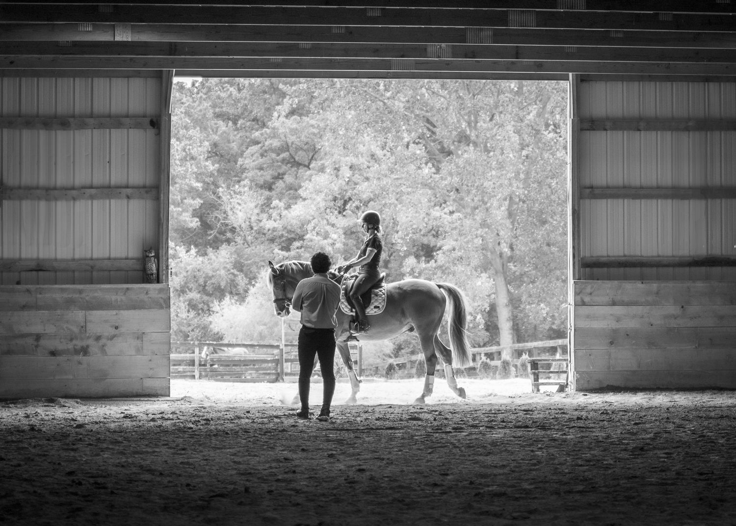 A riding lesson in black and white. Horse, rider and instructor are all framed by a larger open door showing the trees outside.