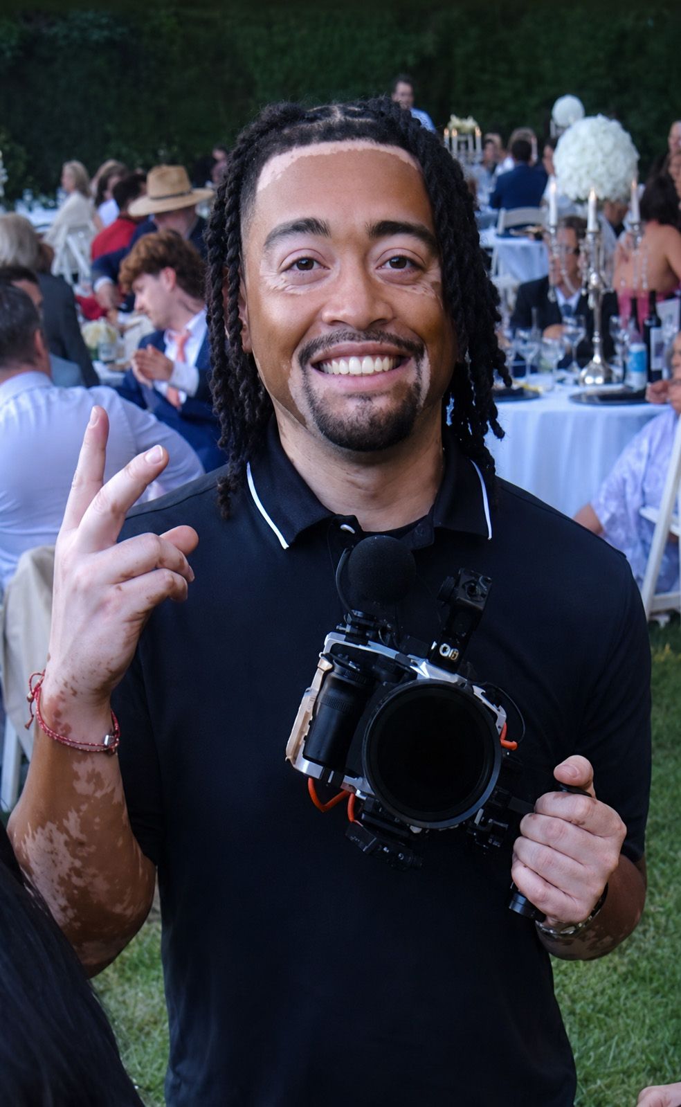 Smiling man with dreadlocks holding a camera and making a peace sign at an outdoor event.