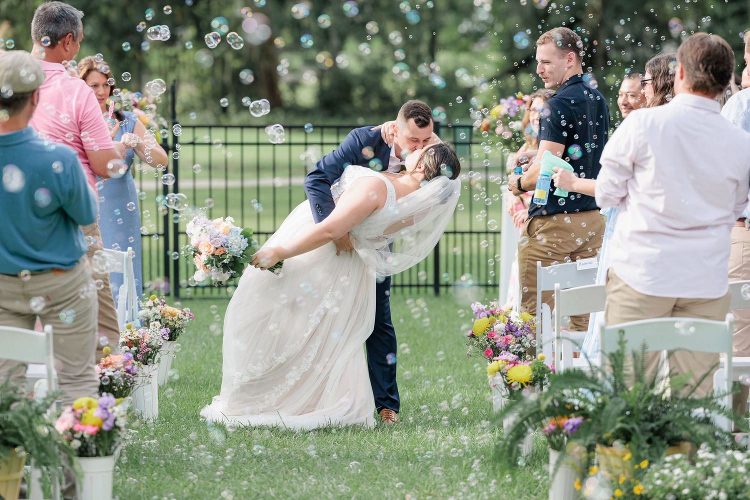 Couple exiting their wedding ceremony and sharing a kiss while the congregation blows bubbles as they exit their backyard wedding.