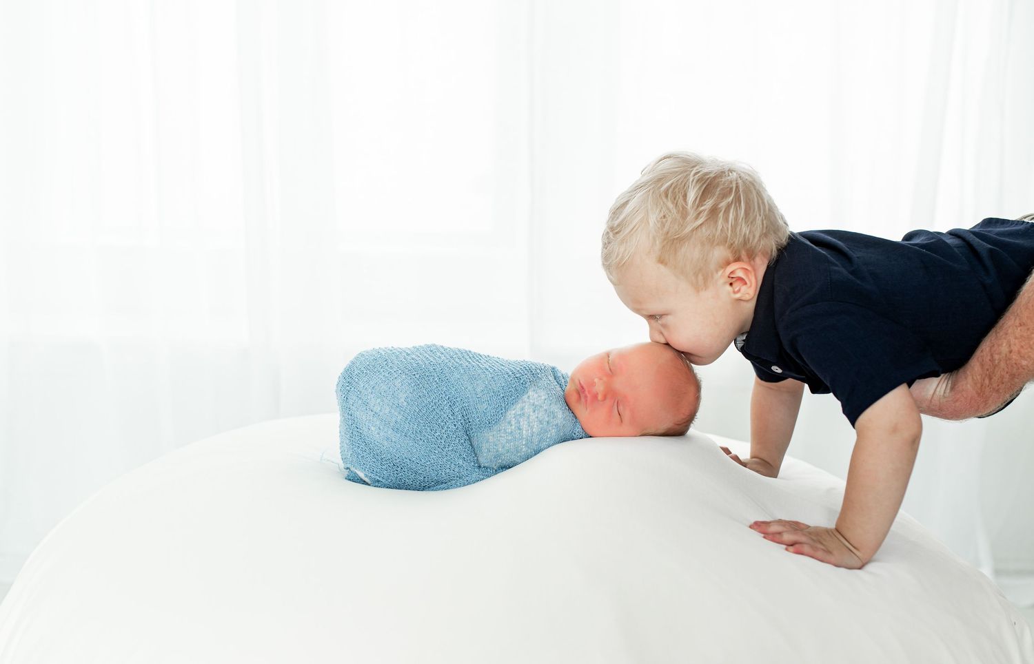 Toddler leaning down to kiss a newborn baby wrapped in blue against a white background.