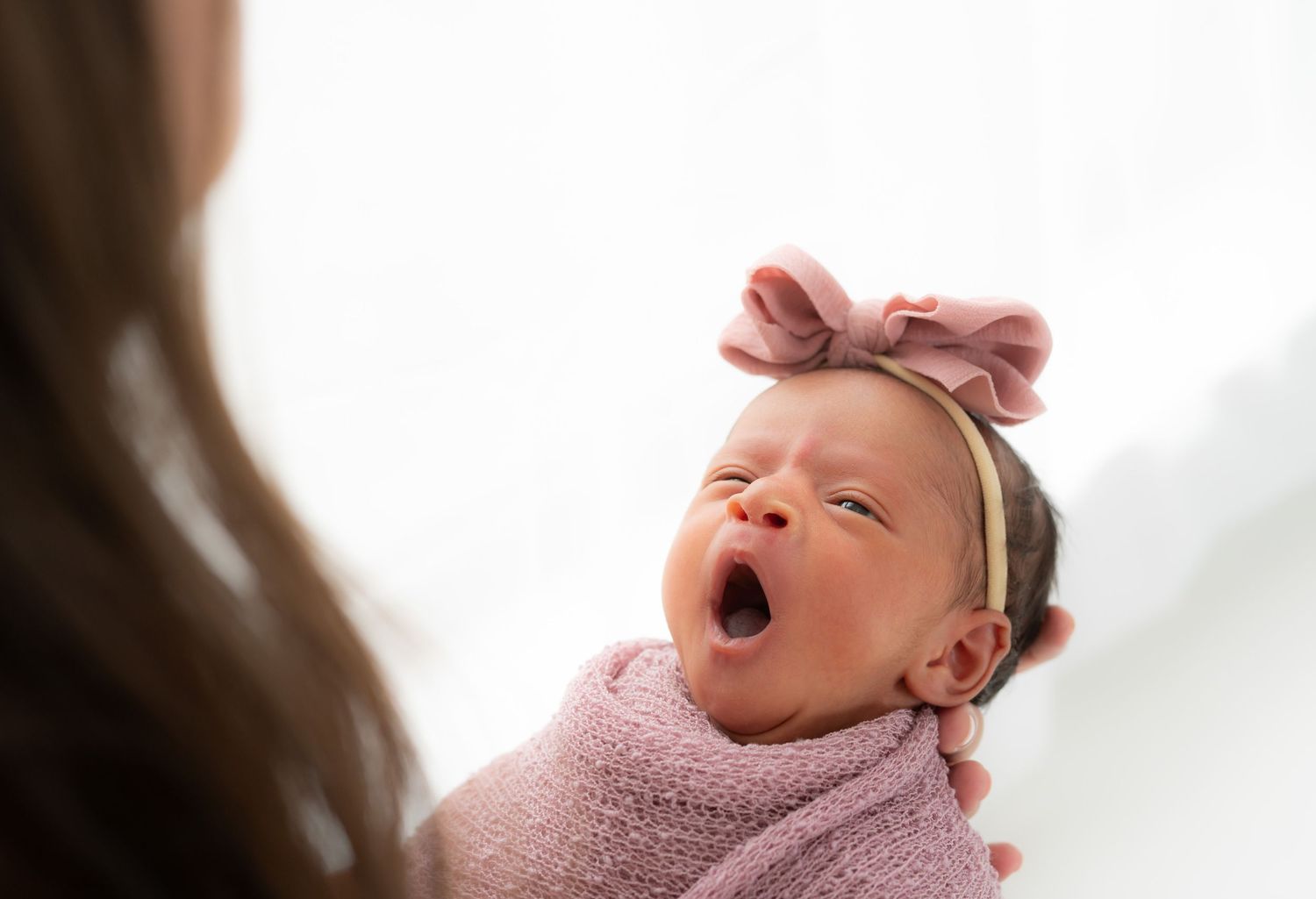 Newborn wearing a pink bow headband yawning while held by her mom.