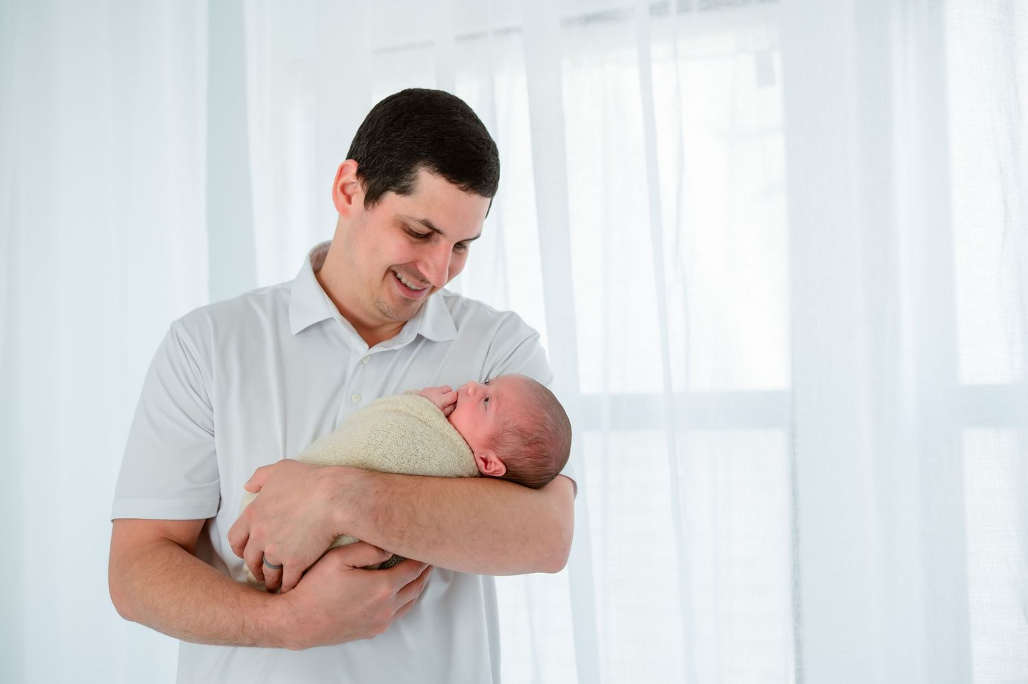 Father looking down at his newborn baby while holding them beside a bright window.