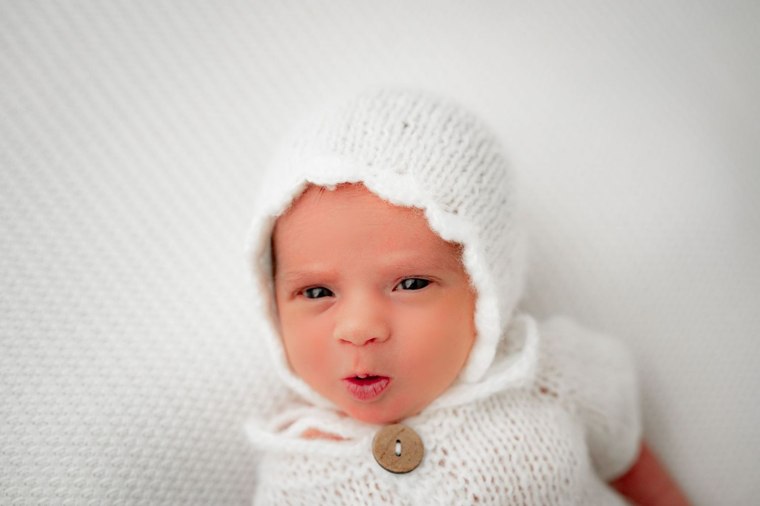 Newborn baby girl in a bonnet and knit romper, looking at the camera during her studio session newborn session in Fulshear, TX.