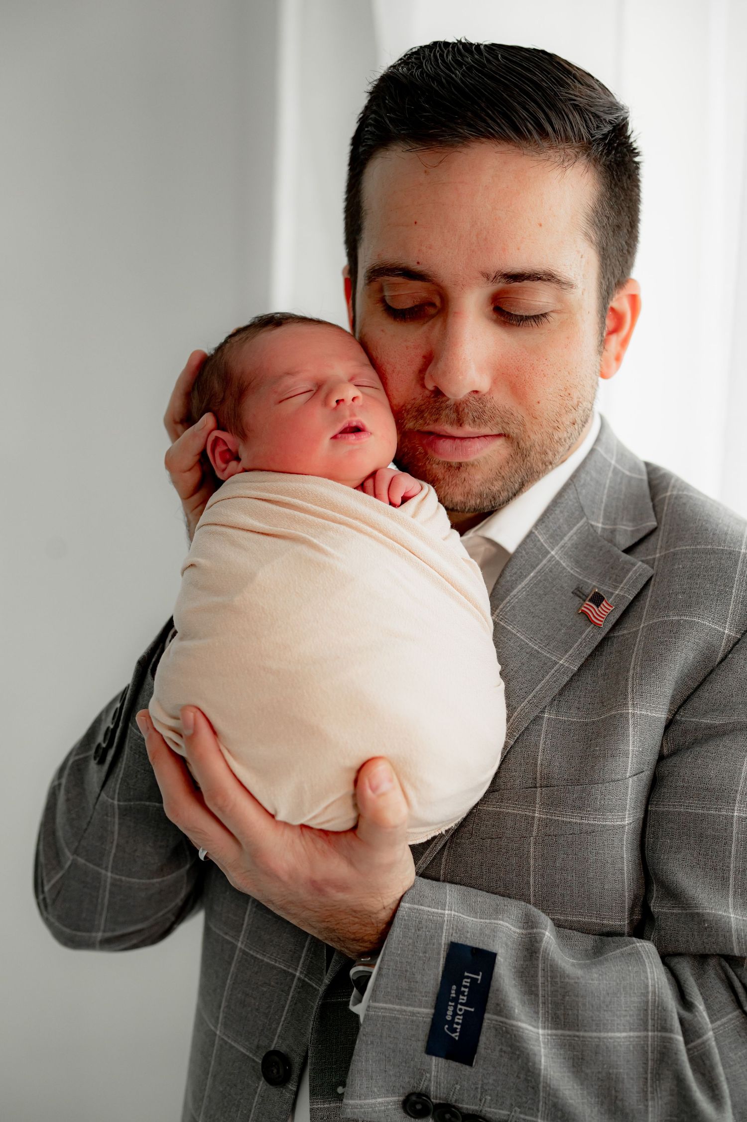 Father in a gray jacket holding a newborn wrapped in cream close to his chest.