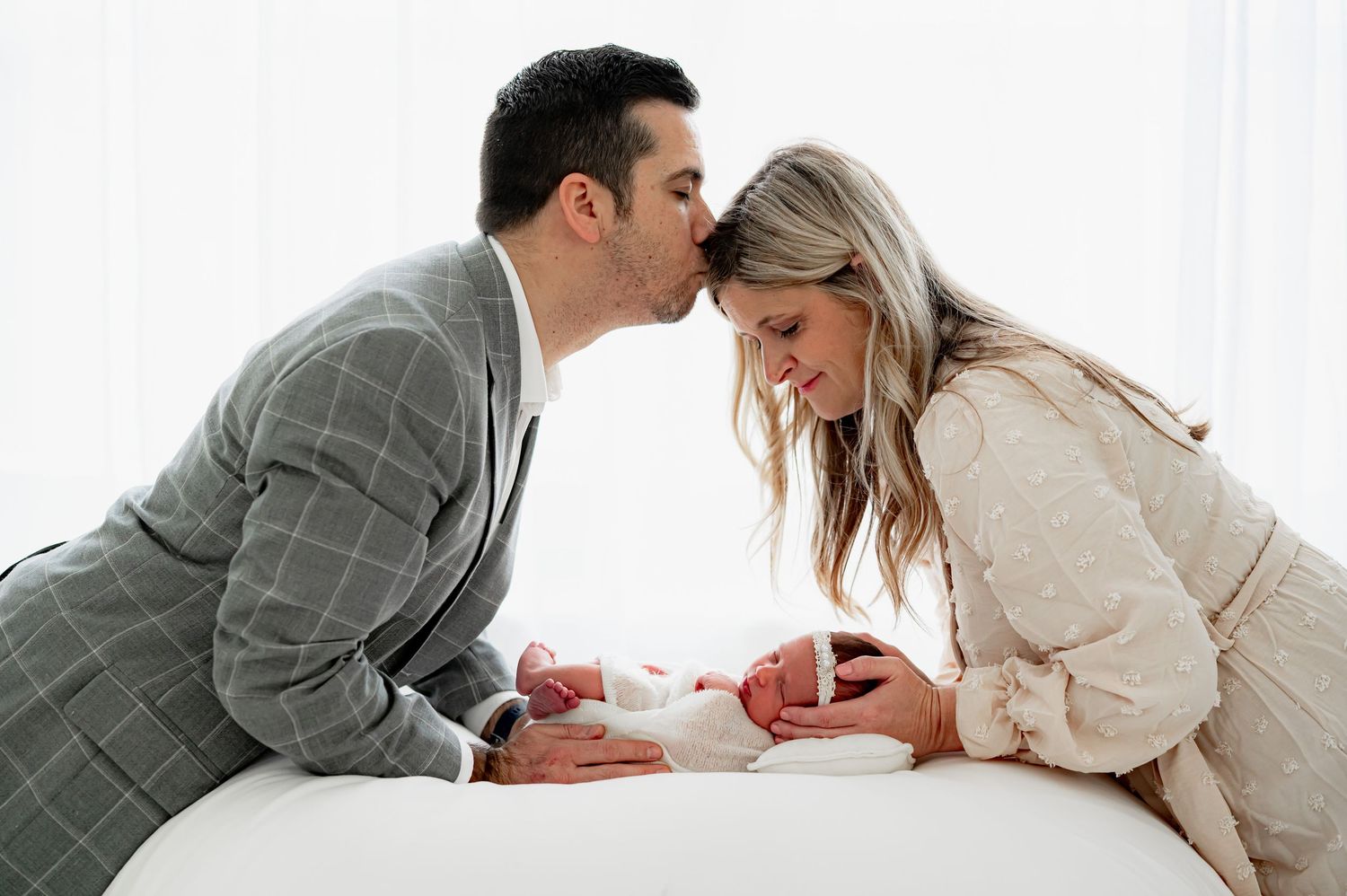 Father kissing his wife while she gazes at their sleeping newborn baby.