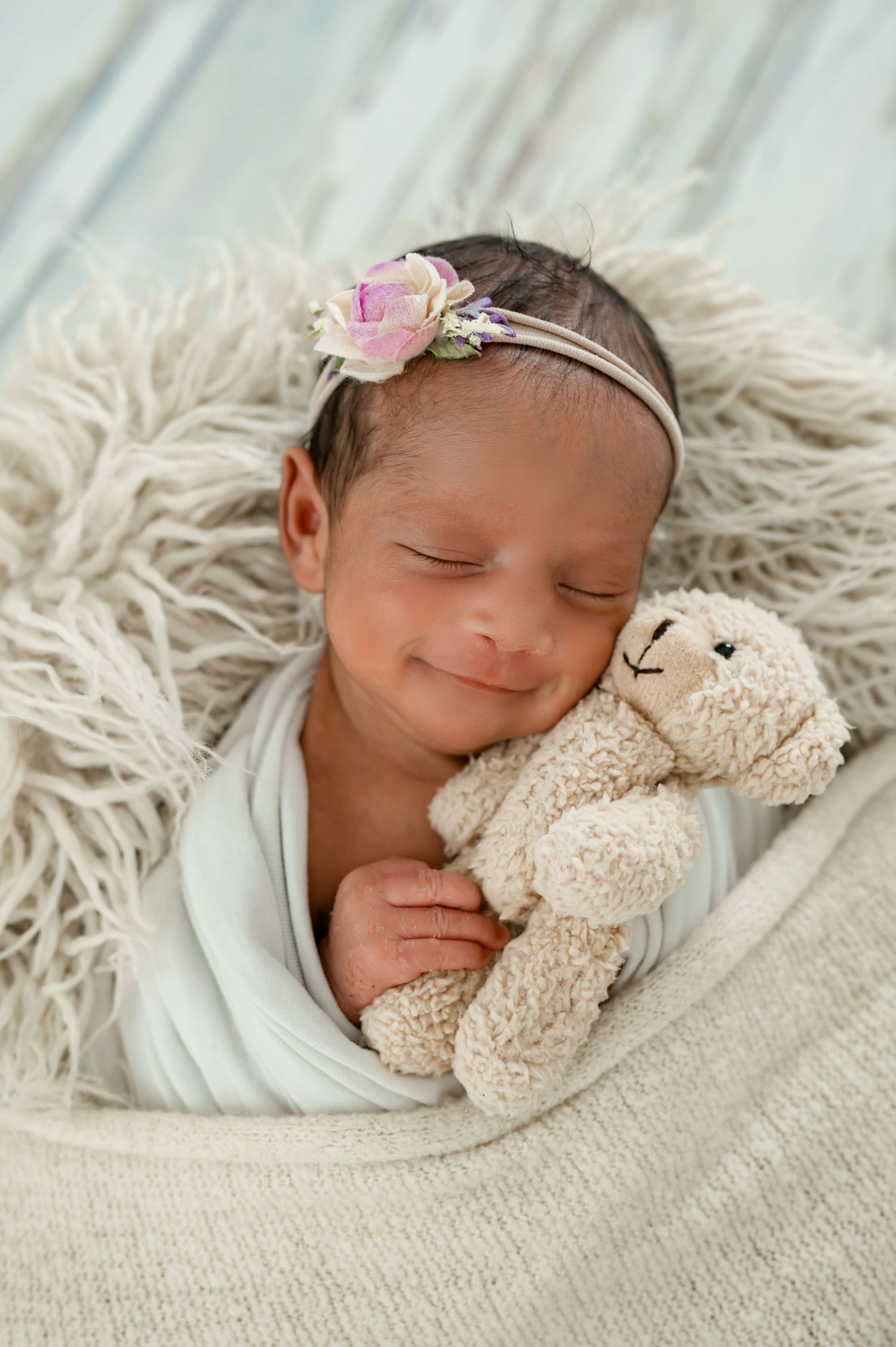 Sleeping newborn in a white wrap and headband cuddling a small stuffed animal while smiling.