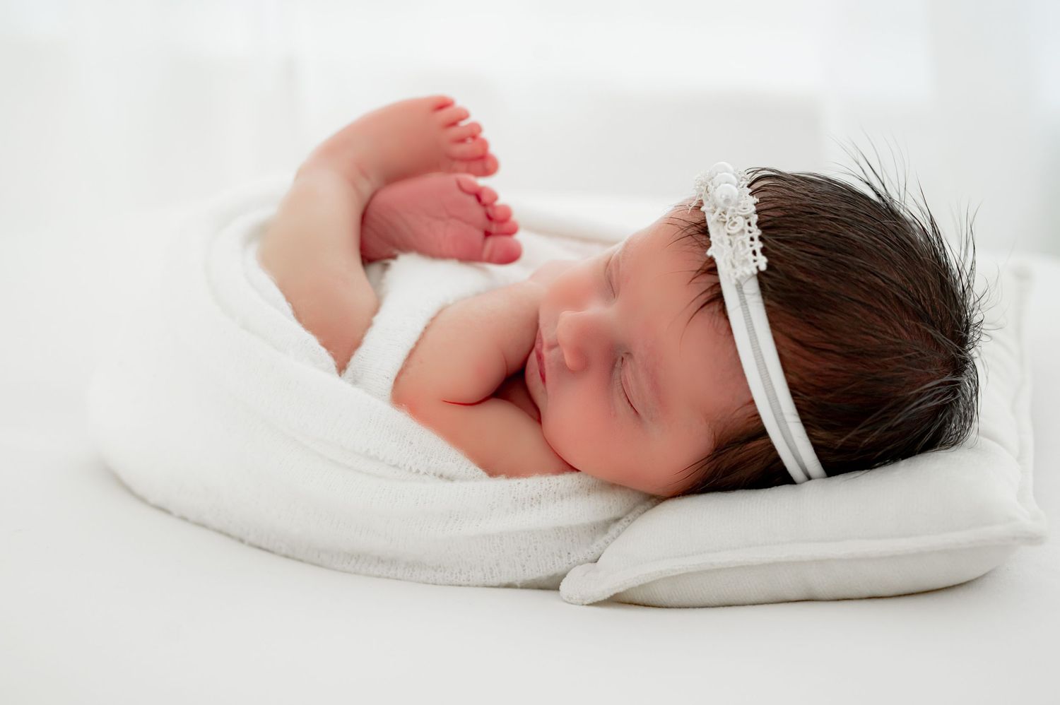 Newborn baby with dark hair and white headband on a white pillow.