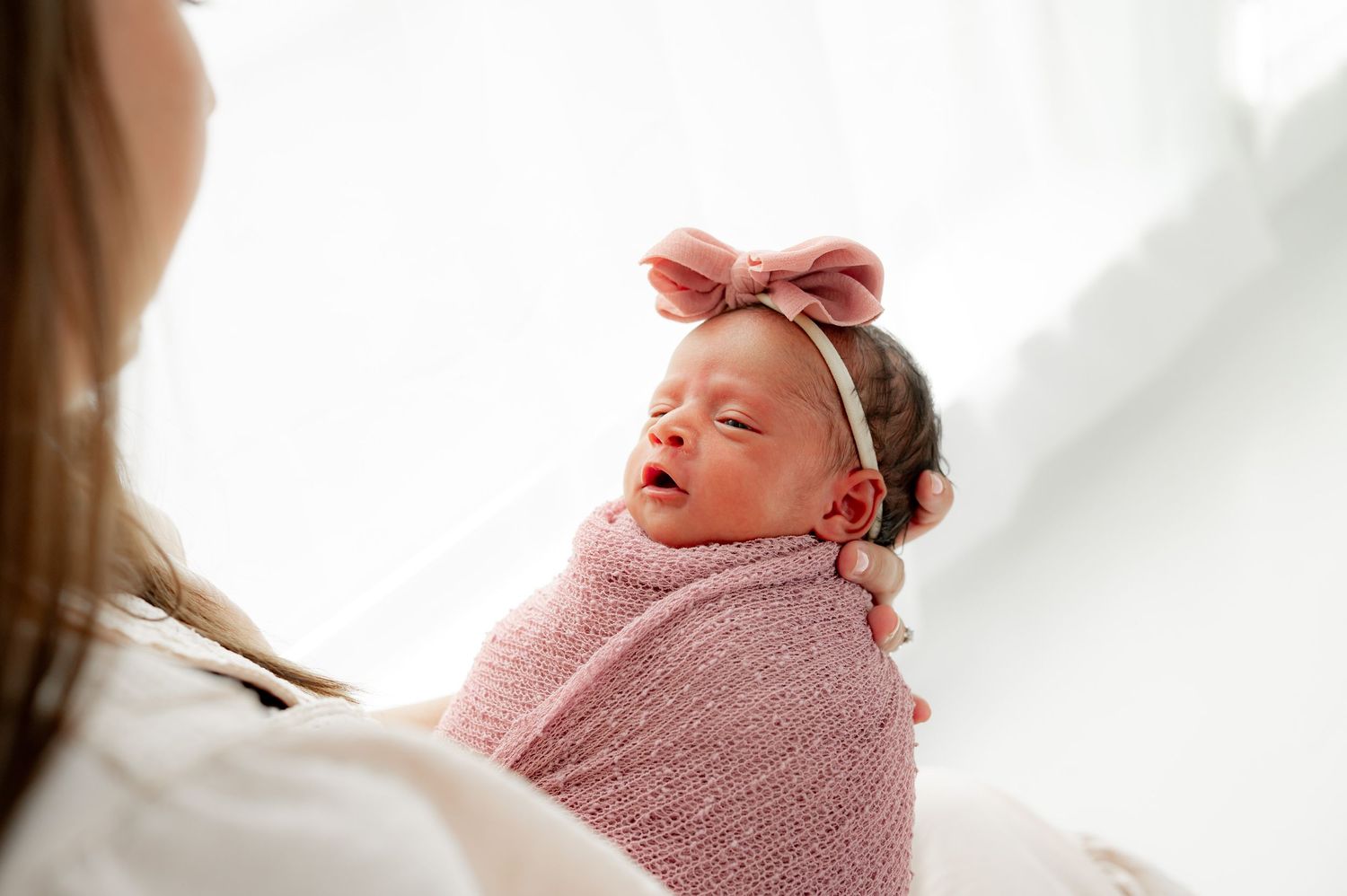 Newborn baby girl wearing a pink bow and wrapped in a pink wrap, gazing at her mama during her newborn studio session in Fulshear, TX.