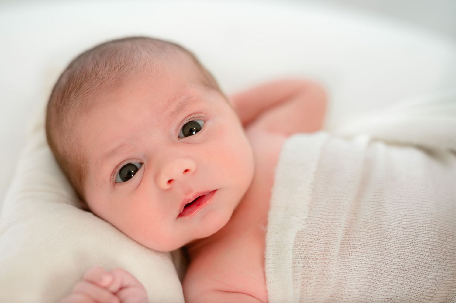 Awake newborn baby wrapped in a blanket and resting on a white pillow.