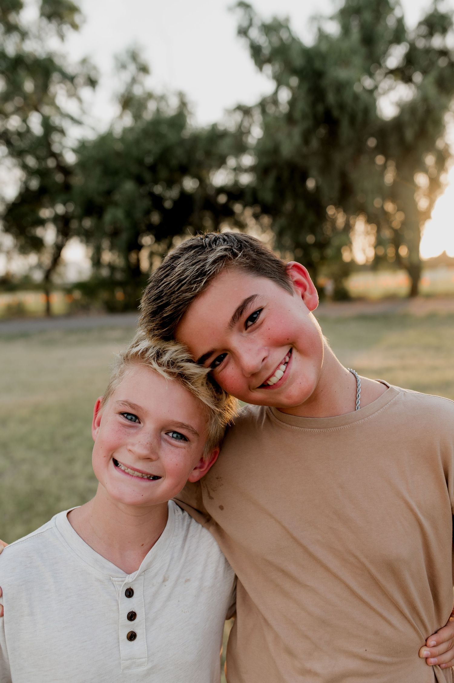 Two smiling boys pose together outdoors during golden hour, with lush green trees in the background.