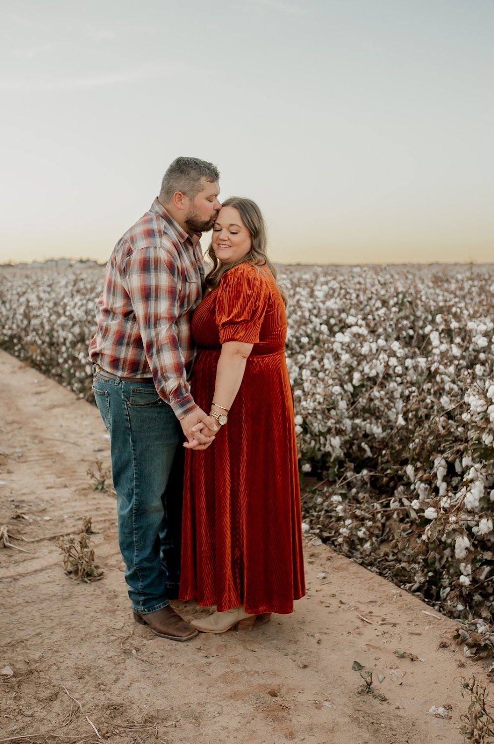 Couple embracing in a cotton field at sunset, woman in red velvet dress, man in plaid shirt and jeans.