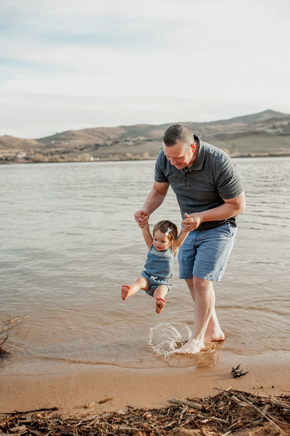 A father lifts his baby girl while splashing in shallow lake water on a sandy beach with hills in the background.