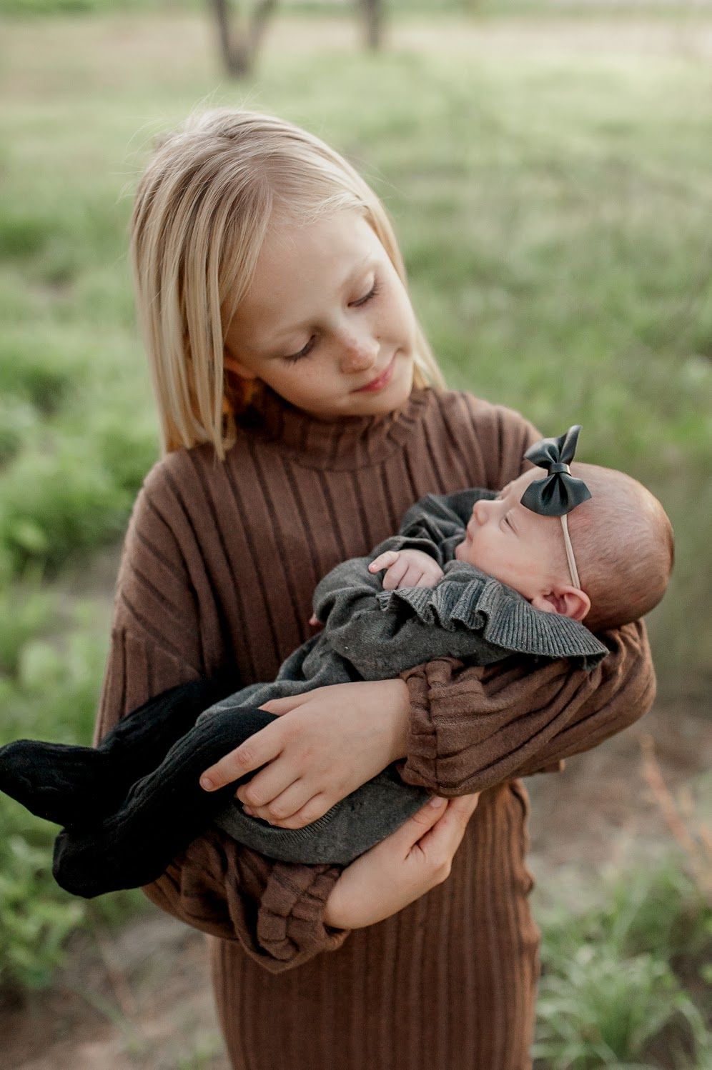 A young blonde girl in a brown ribbed dress tenderly holds a newborn baby dressed in dark gray outdoors.