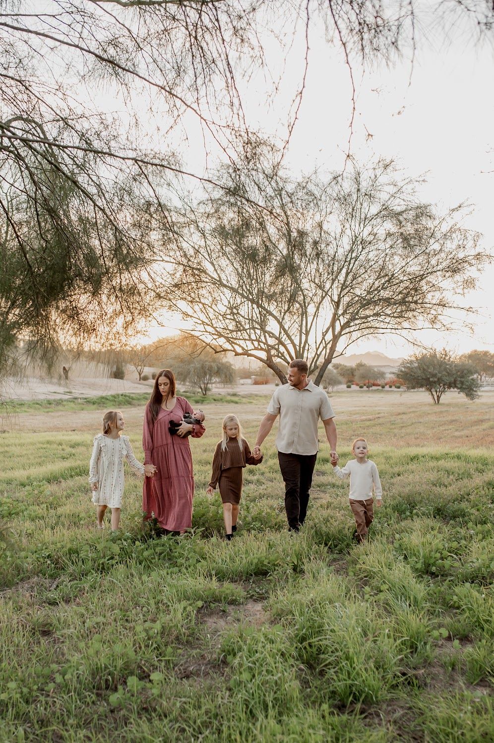 Family of five walking through a sunlit green field at golden hour with trees in the background.