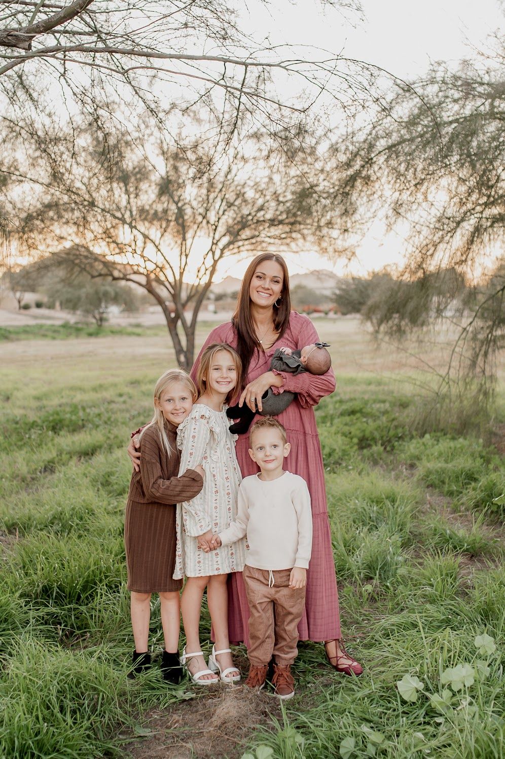 A smiling mother in a pink dress holds a newborn while posing with three young children in a grassy field at sunset.