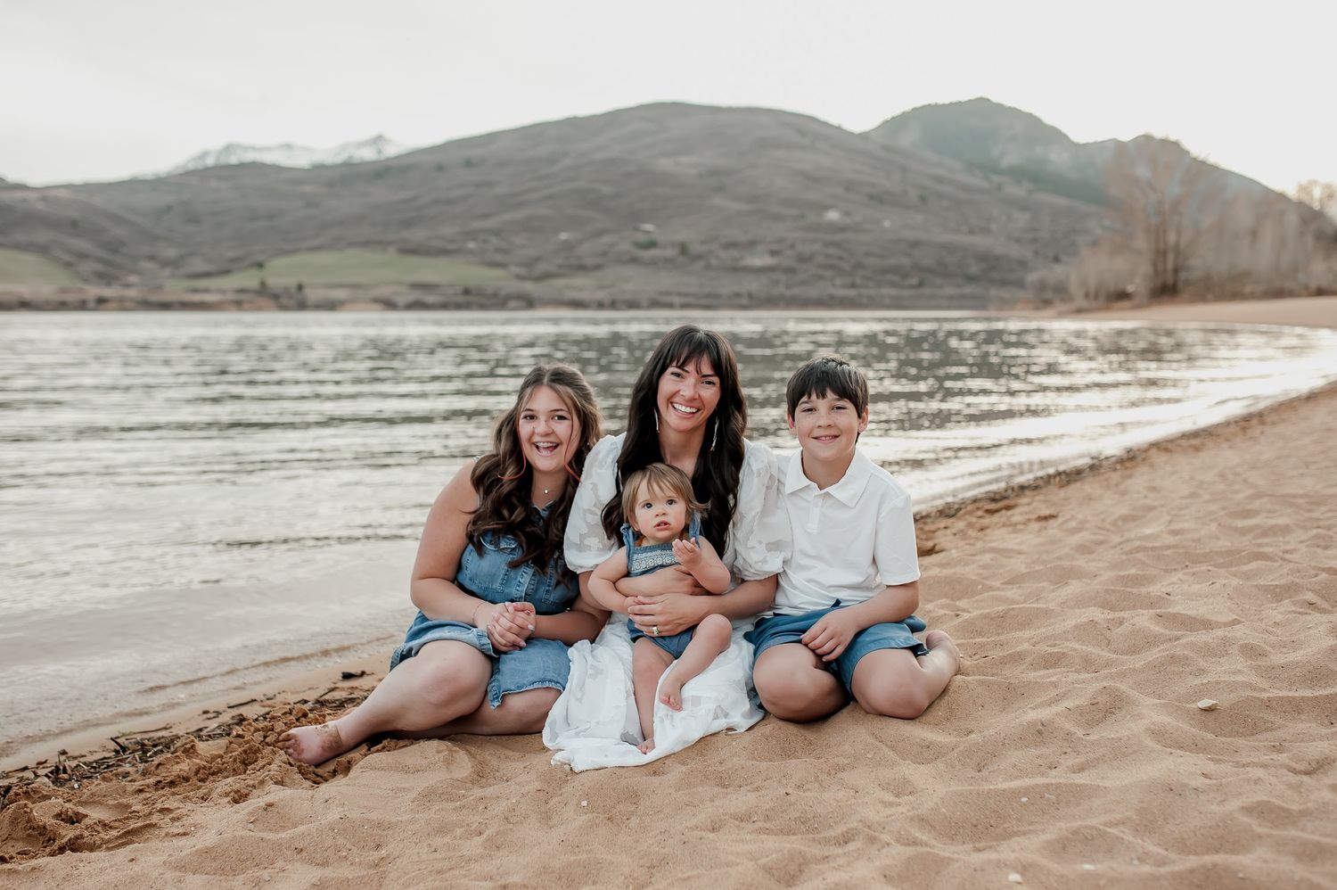Family of four sitting on sandy beach shore with mountain lake backdrop, smiling for portrait photo.