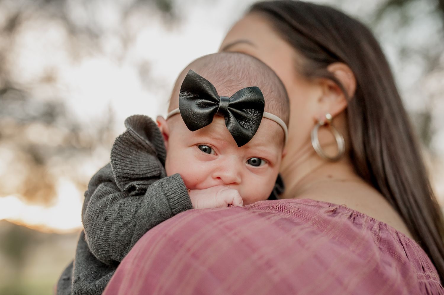 A mother tenderly holds her baby girl wearing a black bow headband and gray outfit in a soft outdoor setting.