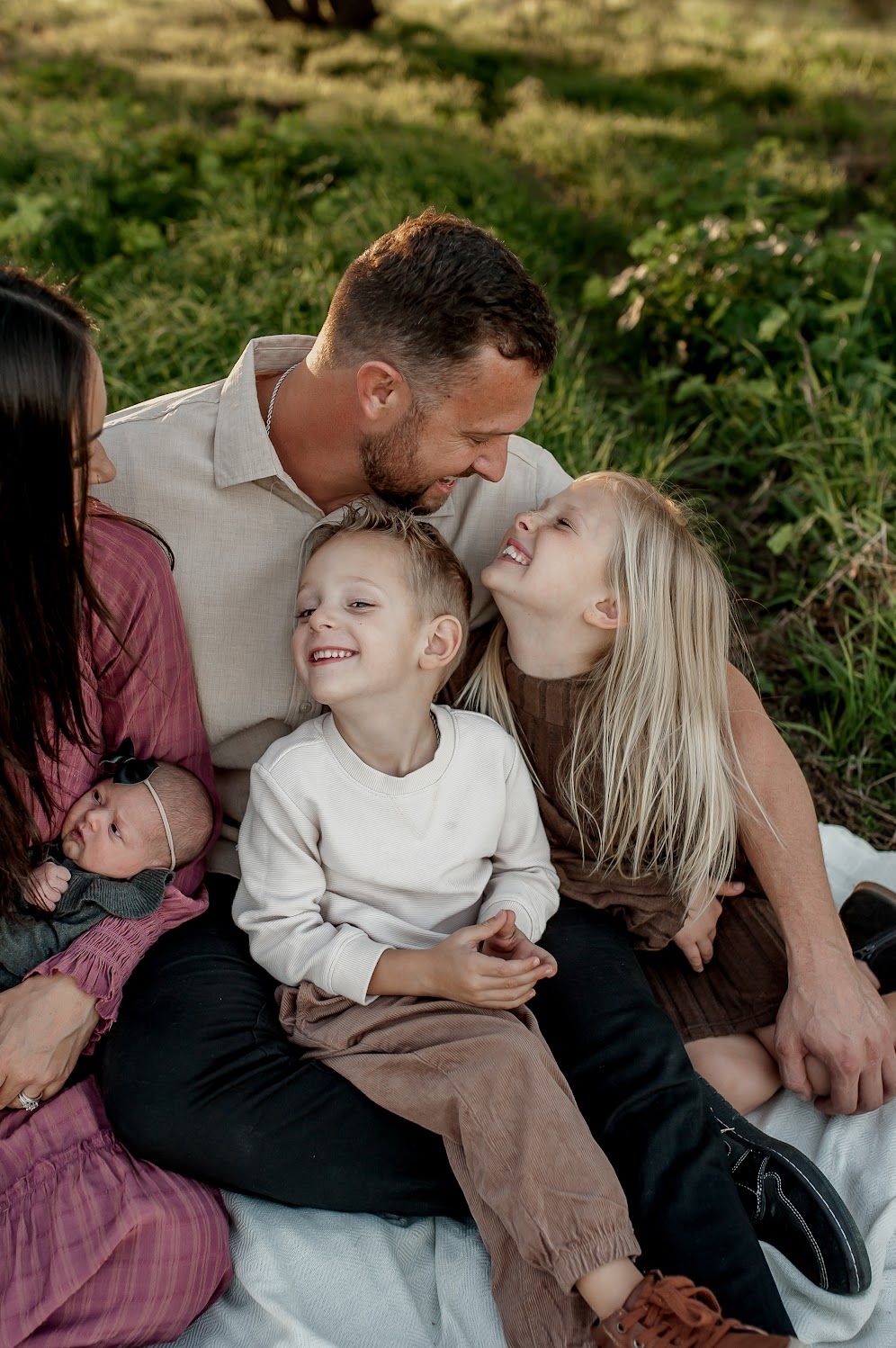 A happy family with two young children and a newborn sits together on a blanket outdoors in lush green grass.