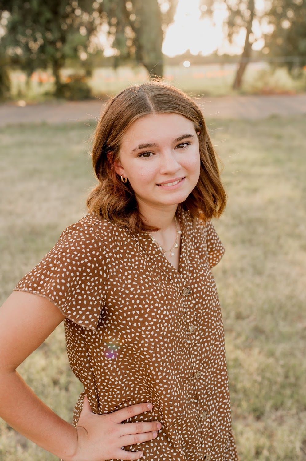 Young woman in brown polka dot dress smiling outdoors at golden hour in a grassy park setting.