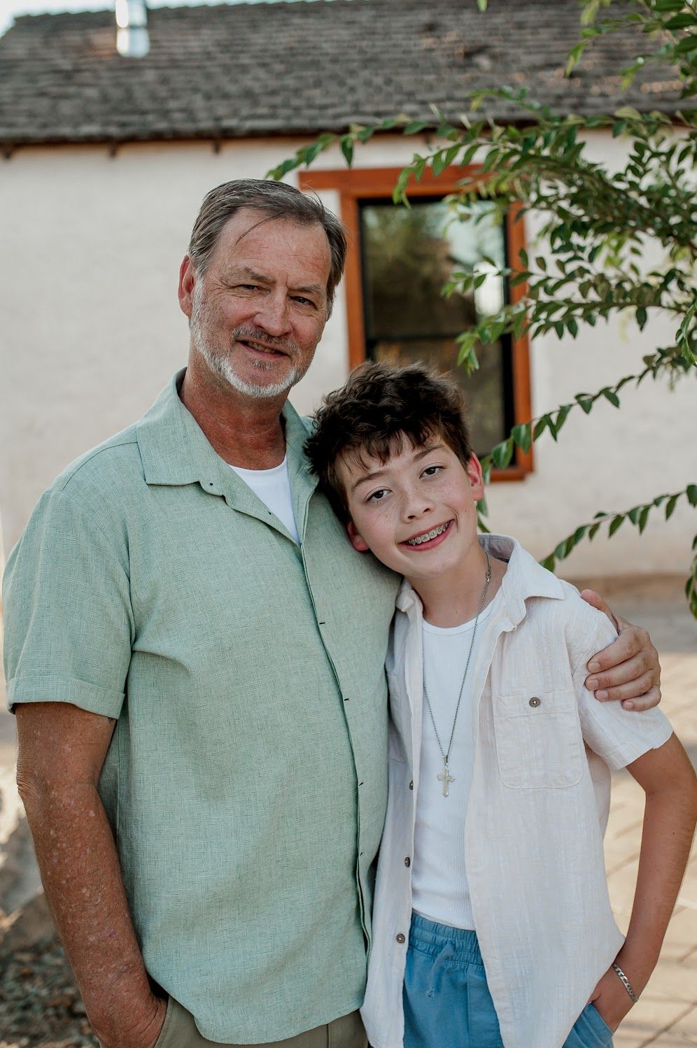 An older man and young boy smile together outdoors near a white building, wearing casual shirts in a warm family portrait.