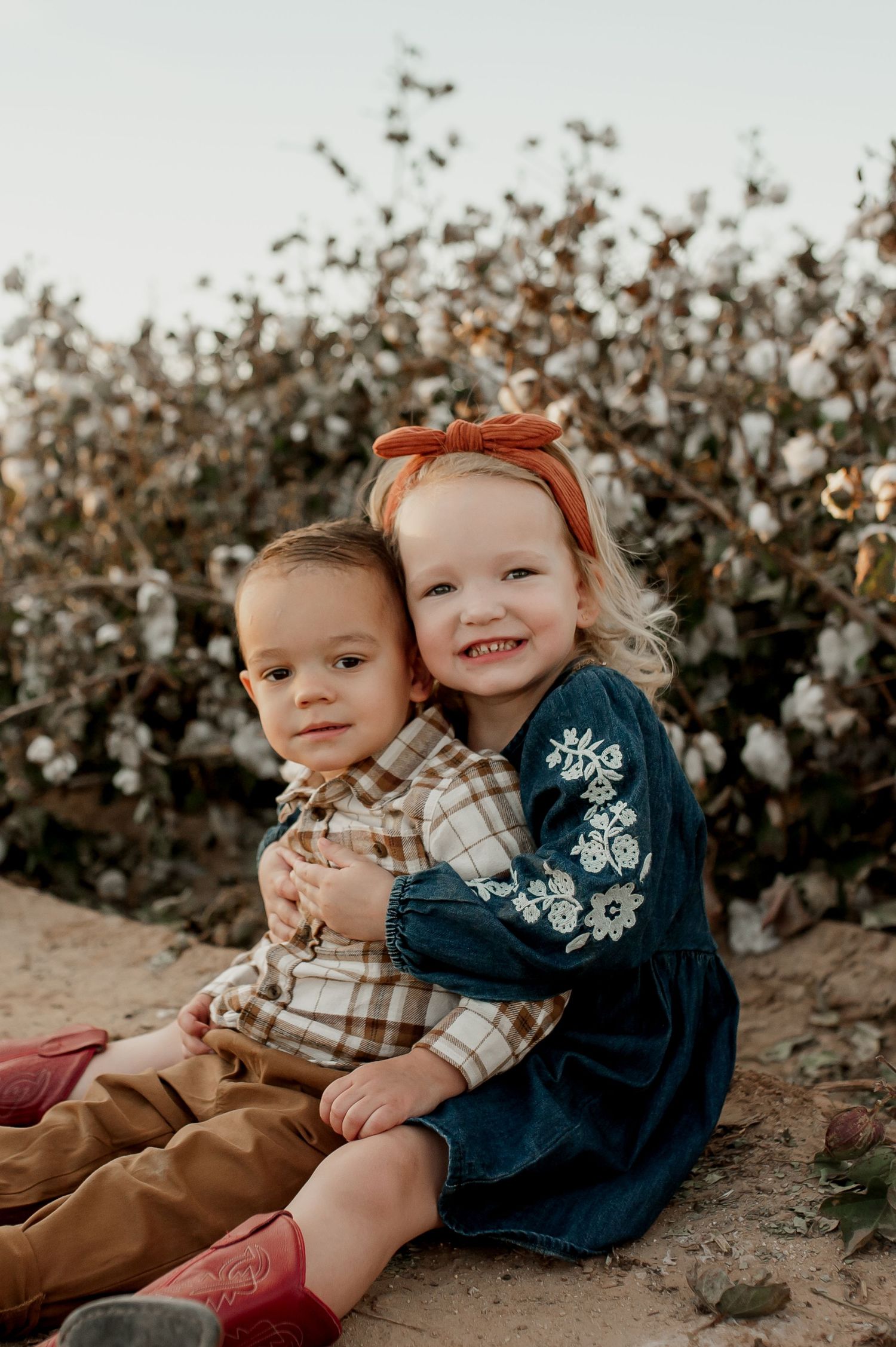 Two young children sit together outdoors near a cotton field, with a girl in a denim dress hugging a boy in a plaid shirt.