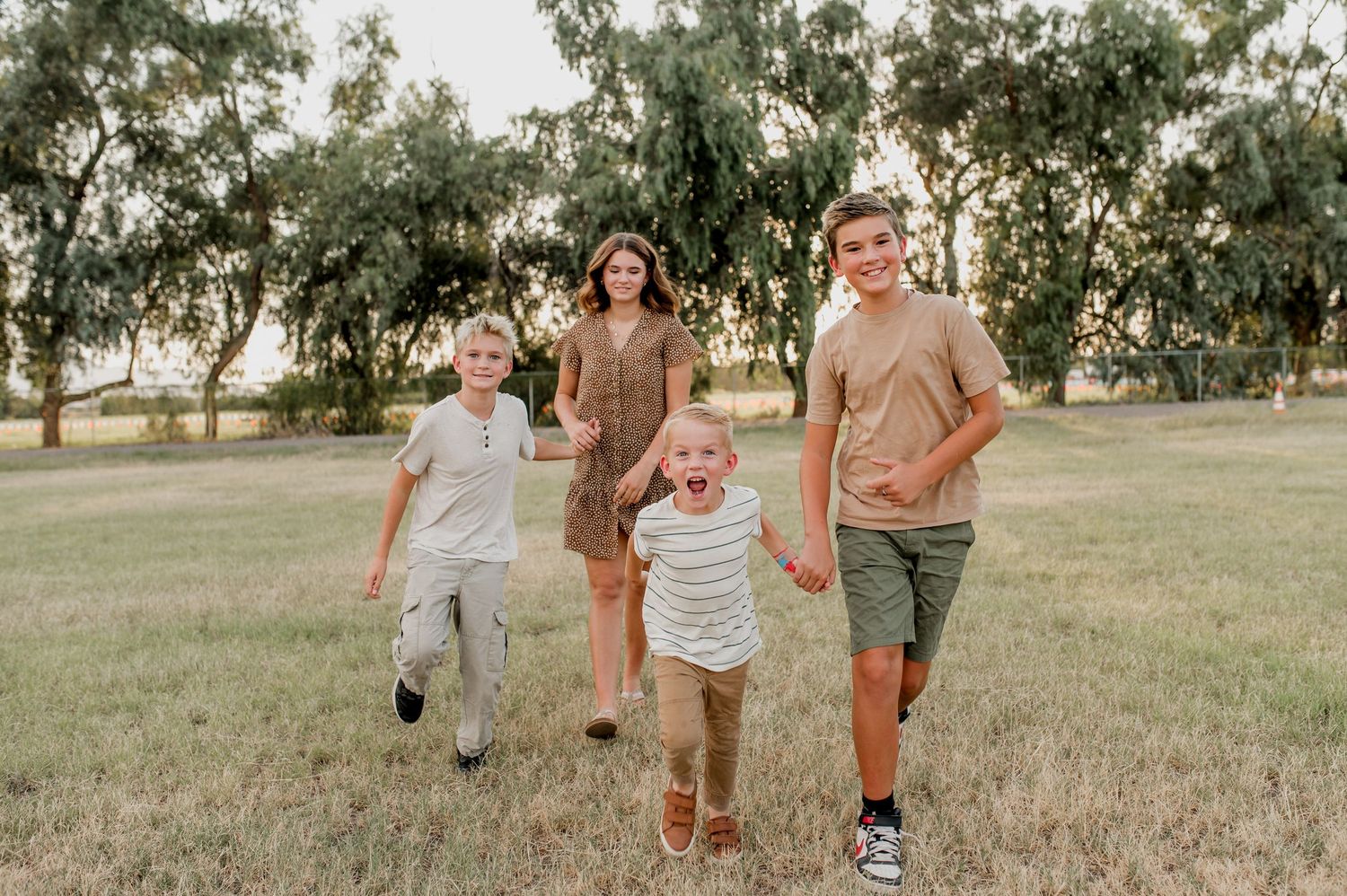 Family of four running and laughing together in a green park with trees on a sunny day.