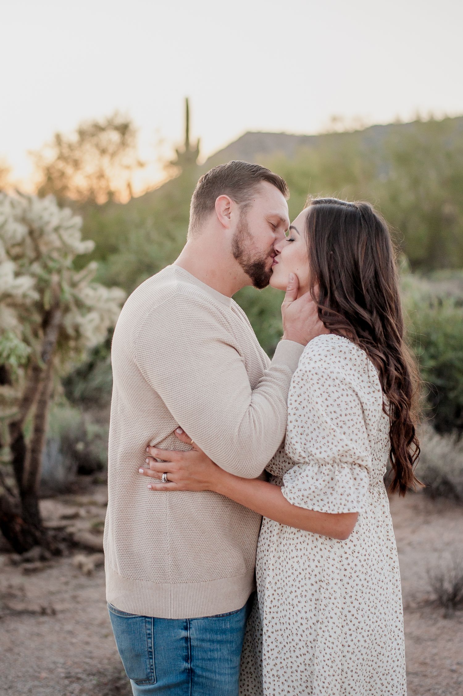 Couple sharing a romantic kiss outdoors in a desert landscape with saguaro cacti at golden hour.