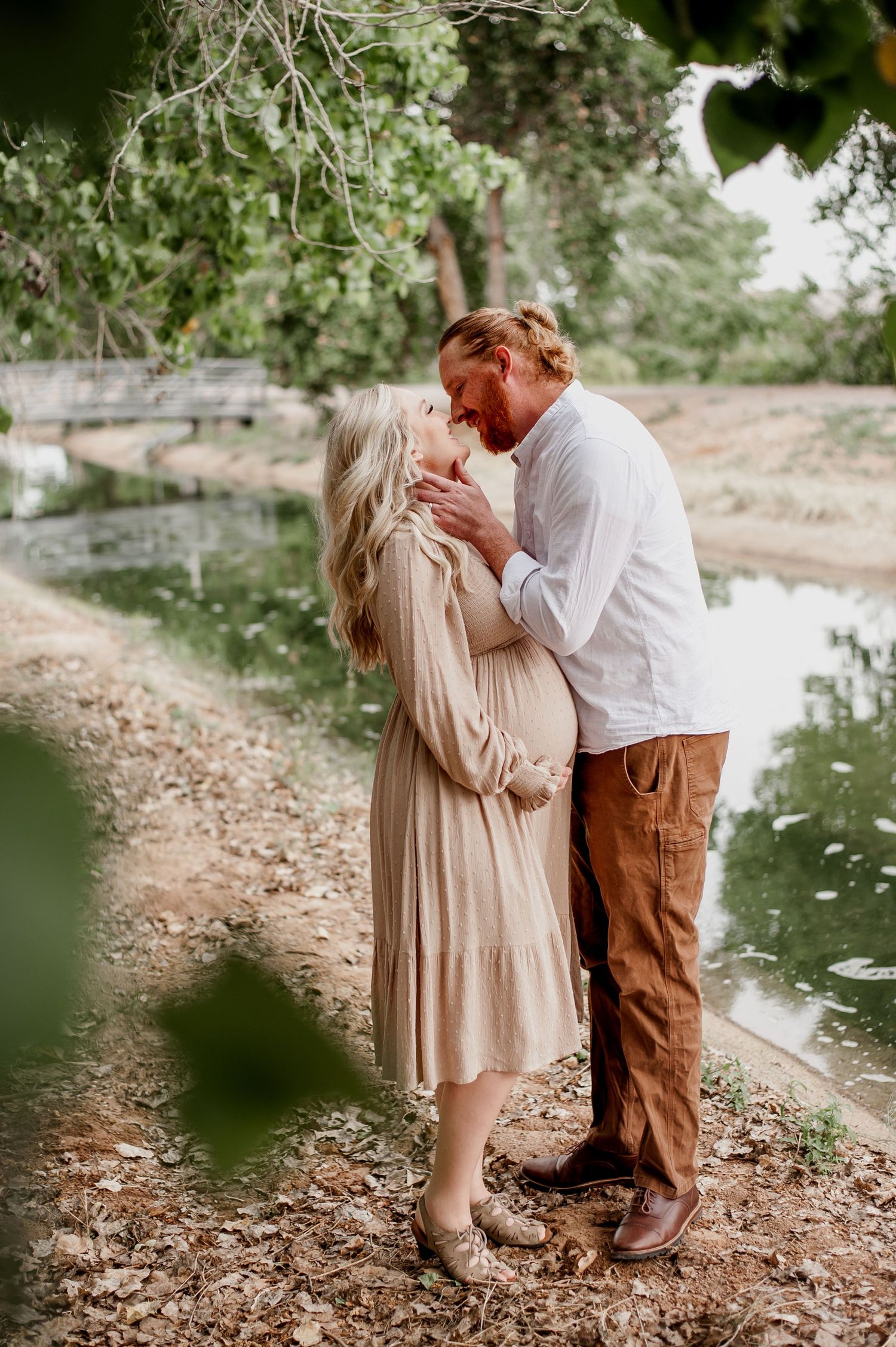 A couple sharing an intimate moment outdoors near a waterway, dressed in neutral earth tones.