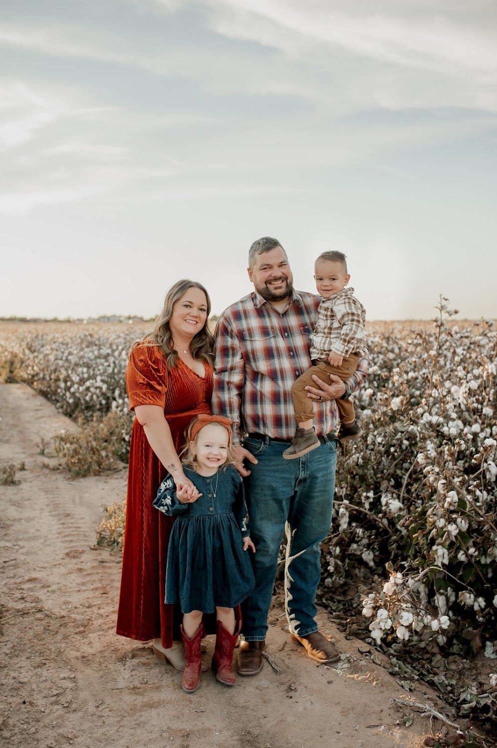 Family of four posing in a cotton field, parents in plaid and red velvet dress, holding two young children.