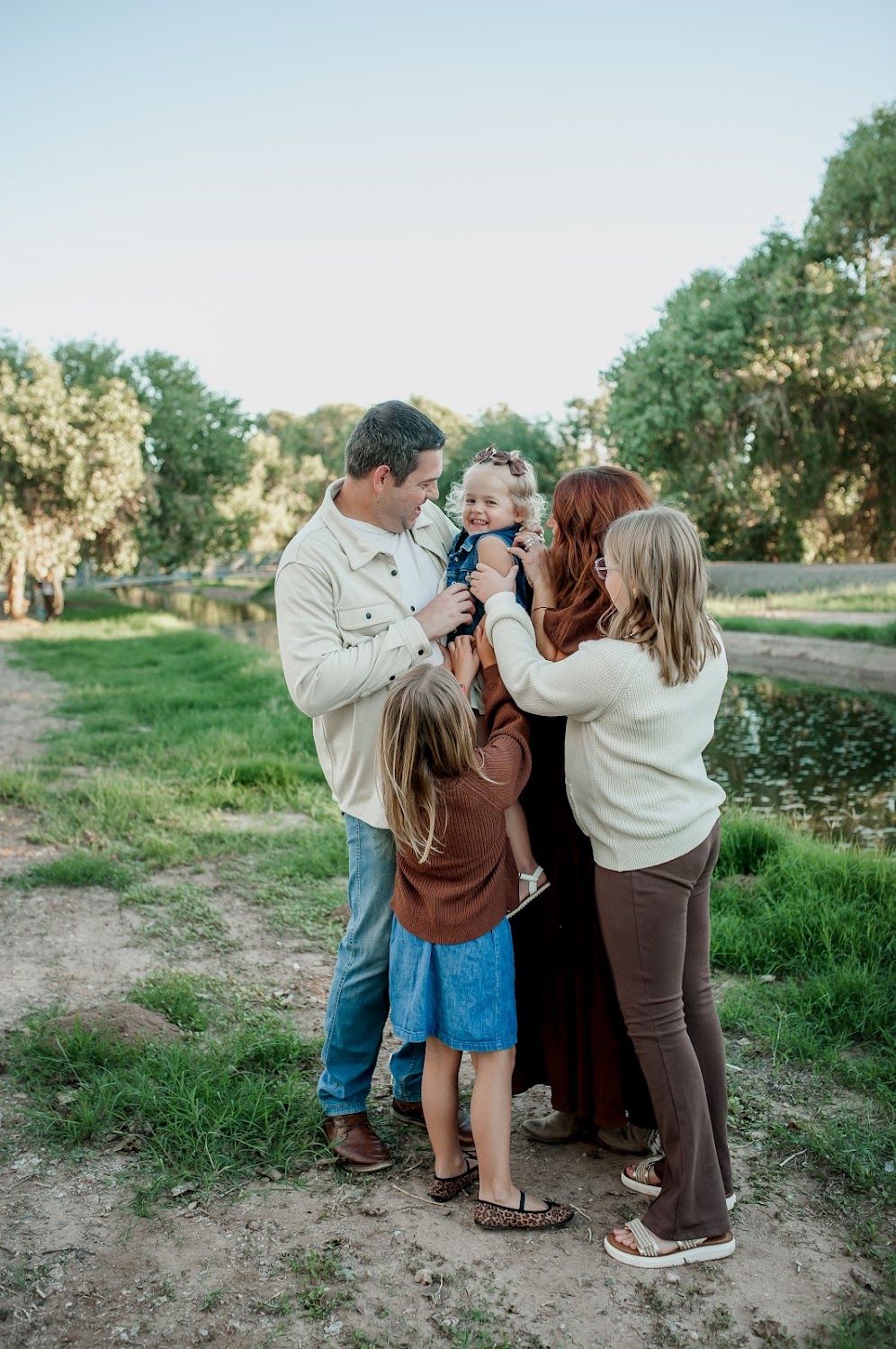 Family of four sharing a joyful moment outdoors near a pond, with lush green trees in the background.
