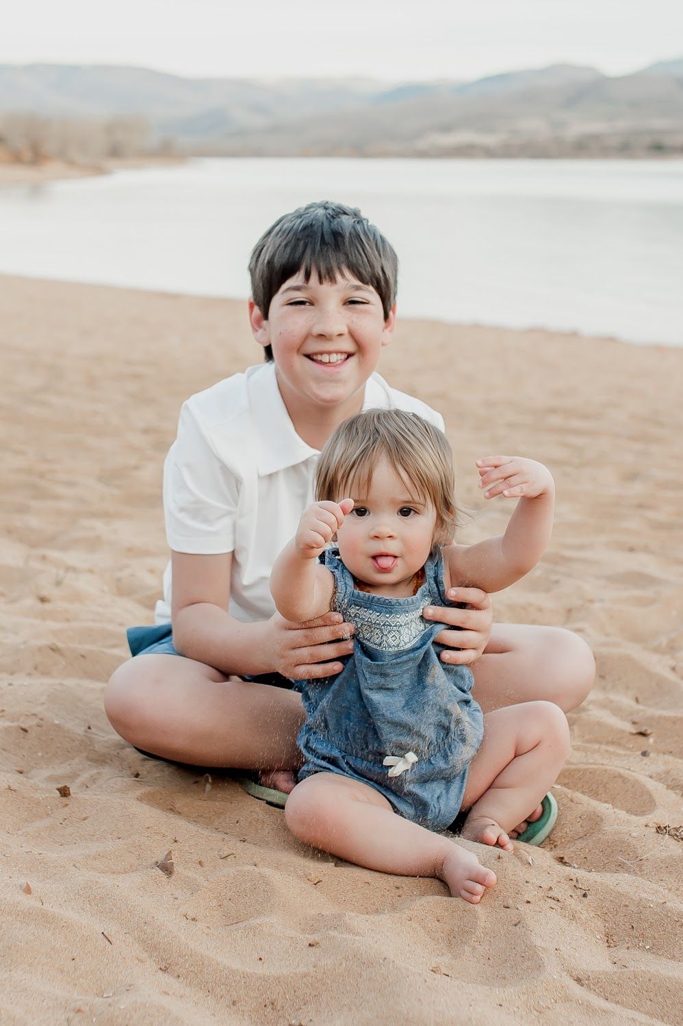Smiling boy in white polo sits cross-legged on sandy beach holding toddler girl in denim overalls near a lake.