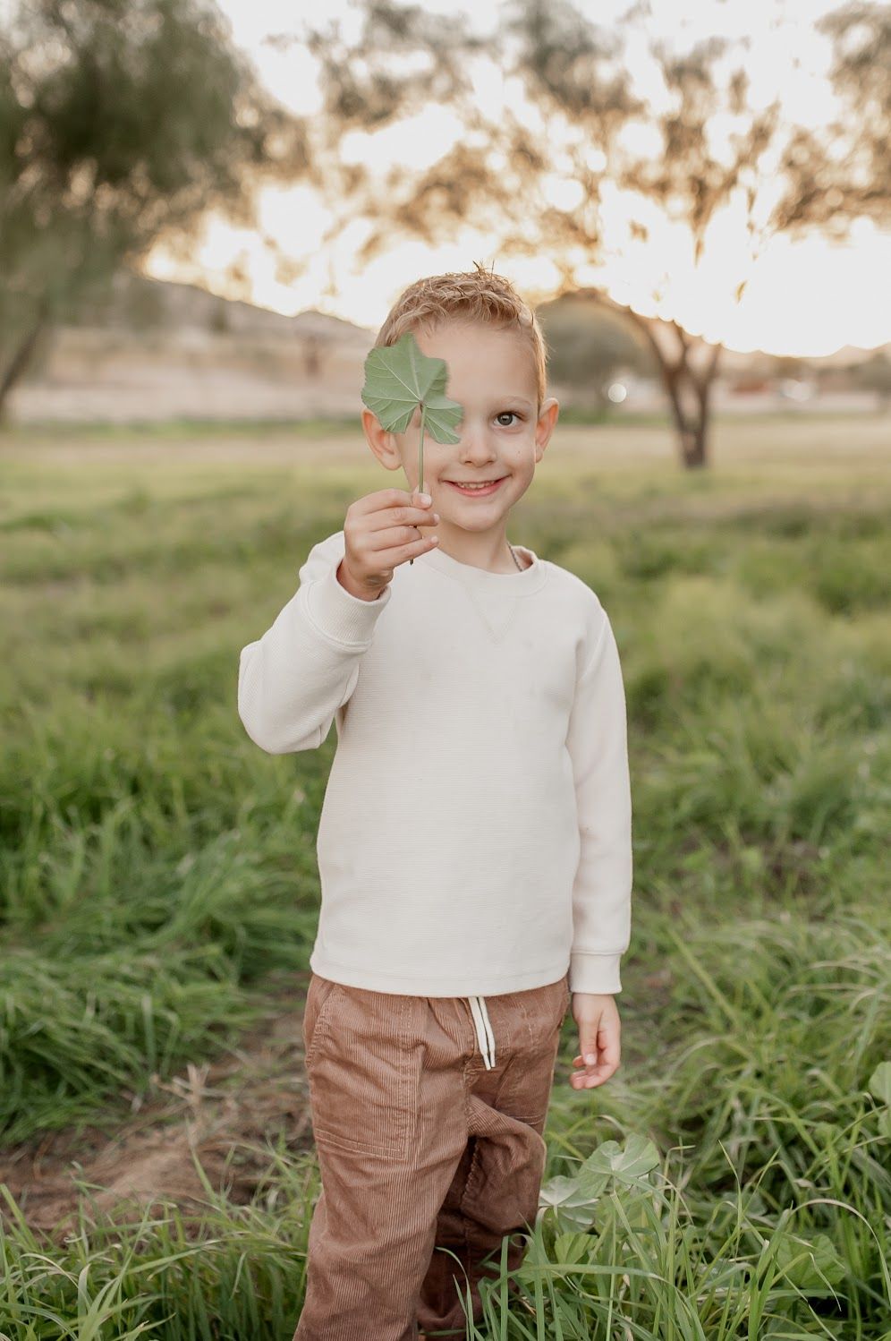 A smiling young boy in a white sweater and brown pants holds a green four-leaf clover in a sunny outdoor field.