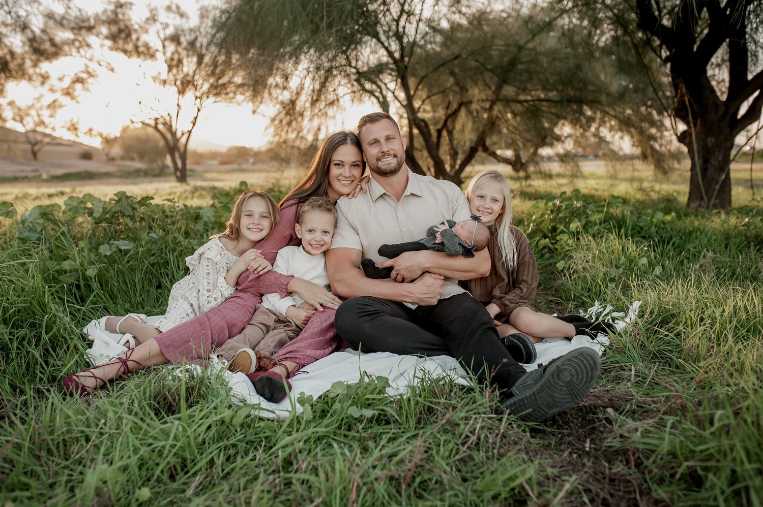 Happy family of five sitting together on a blanket in a lush green field during golden hour sunset.