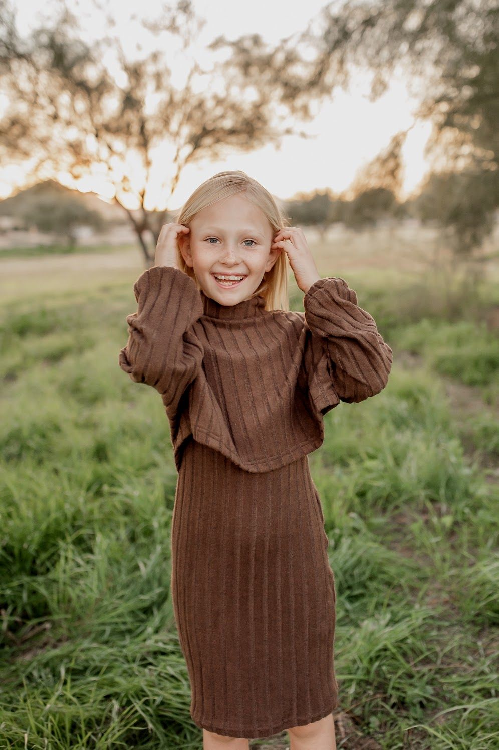 Smiling young girl in brown ribbed turtleneck dress standing in green field at golden hour.