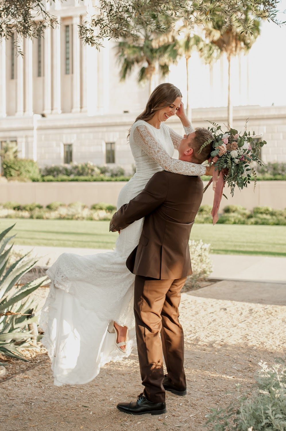 Groom lifts bride in white lace dress outdoors near elegant building, sharing romantic moment with floral bouquet.