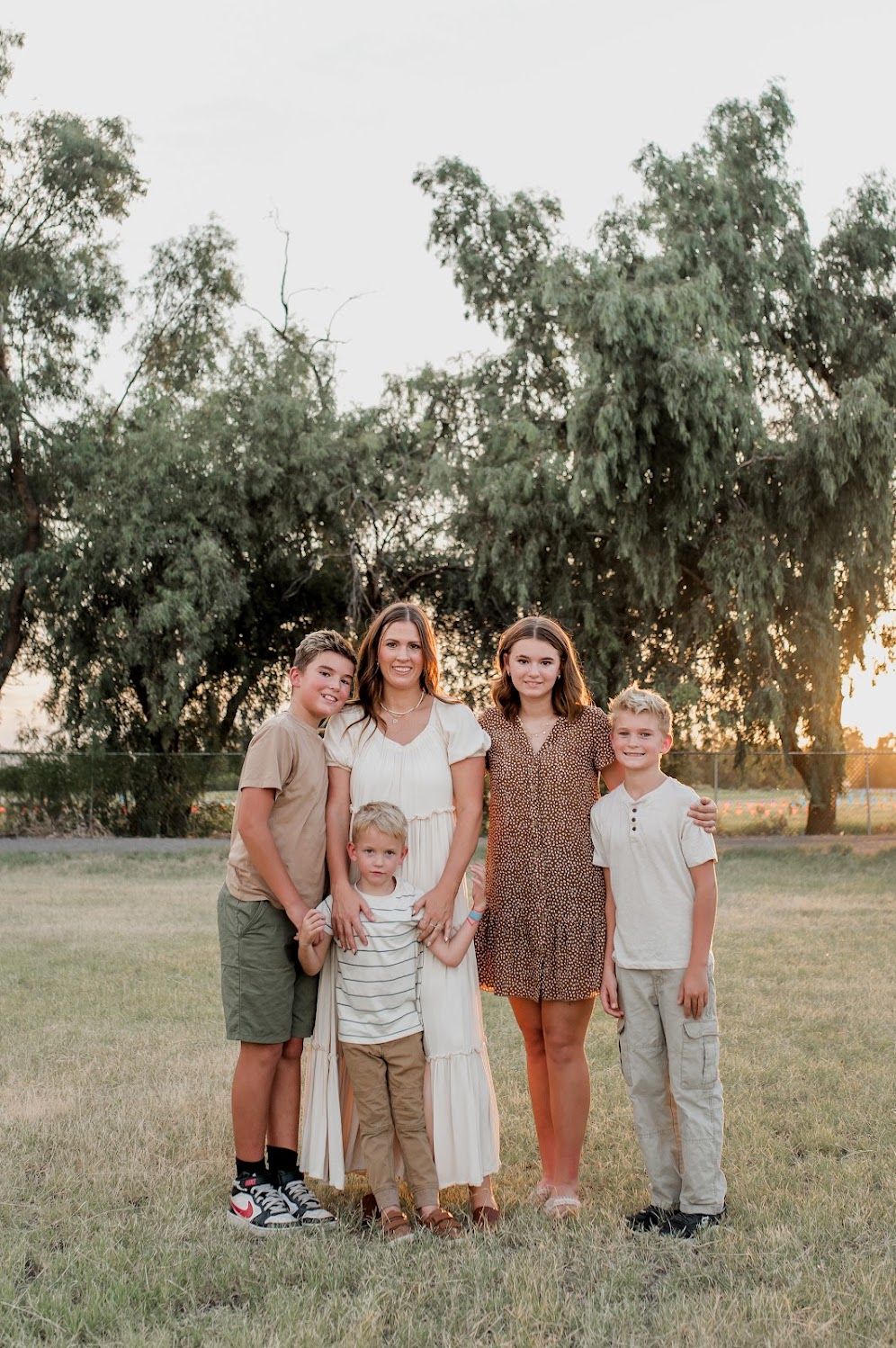 A family of five poses outdoors at golden hour in a grassy field with trees in the background.