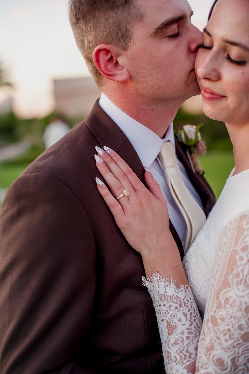 Groom in brown suit kisses bride wearing lace dress and diamond ring during golden hour wedding portrait.
