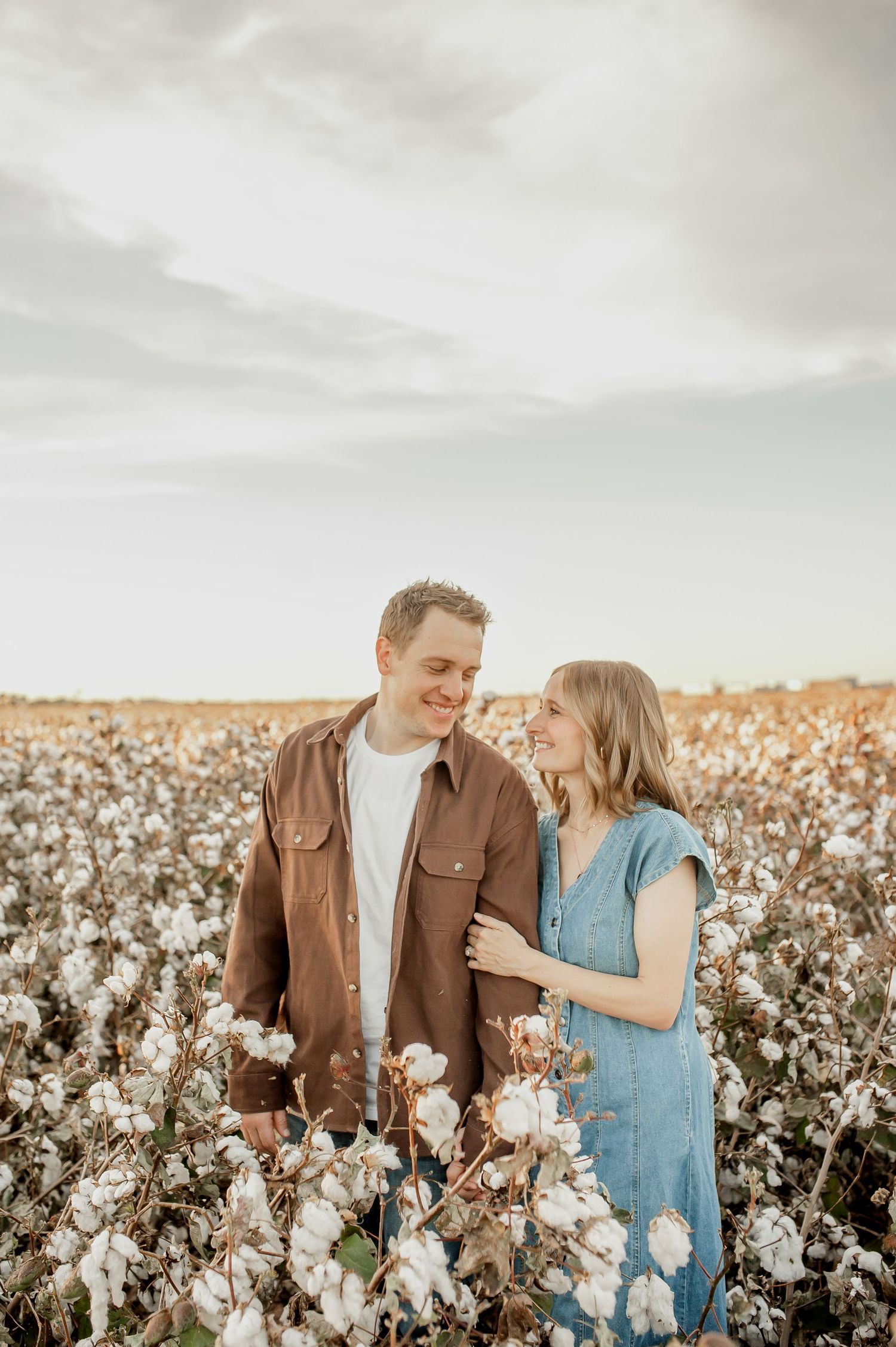 Couple smiling at each other in a cotton field, woman in blue dress, man in brown jacket, cloudy sky backdrop.