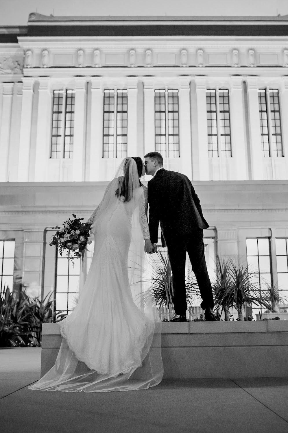 Black and white photo of bride and groom holding hands facing a grand building on their wedding day.